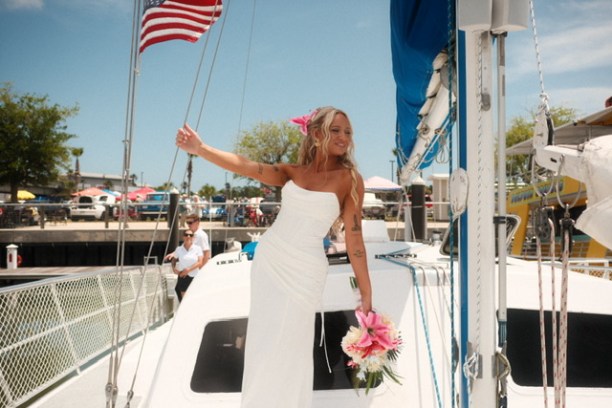 Bride in white gown holding flowers stands on a boat, with American flag waving above.