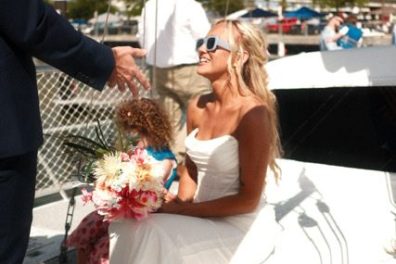 Bride in sunglasses kneeling on boat deck with bouquet, person in suit nearby.